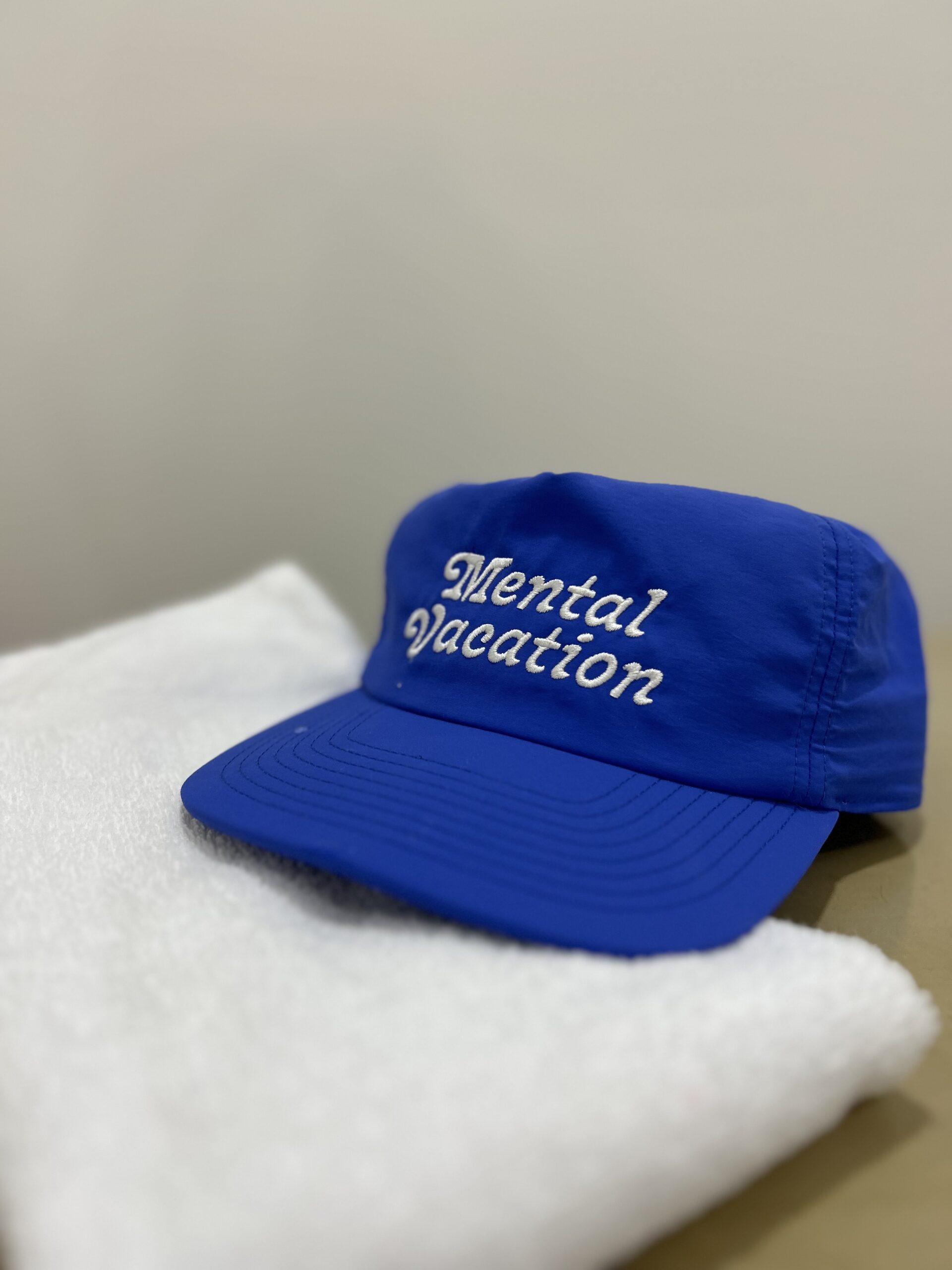 A blue baseball cap faces head-on with the words "Mental Vacation" in an italics serif font, its brim sitting on a white, folded towel
