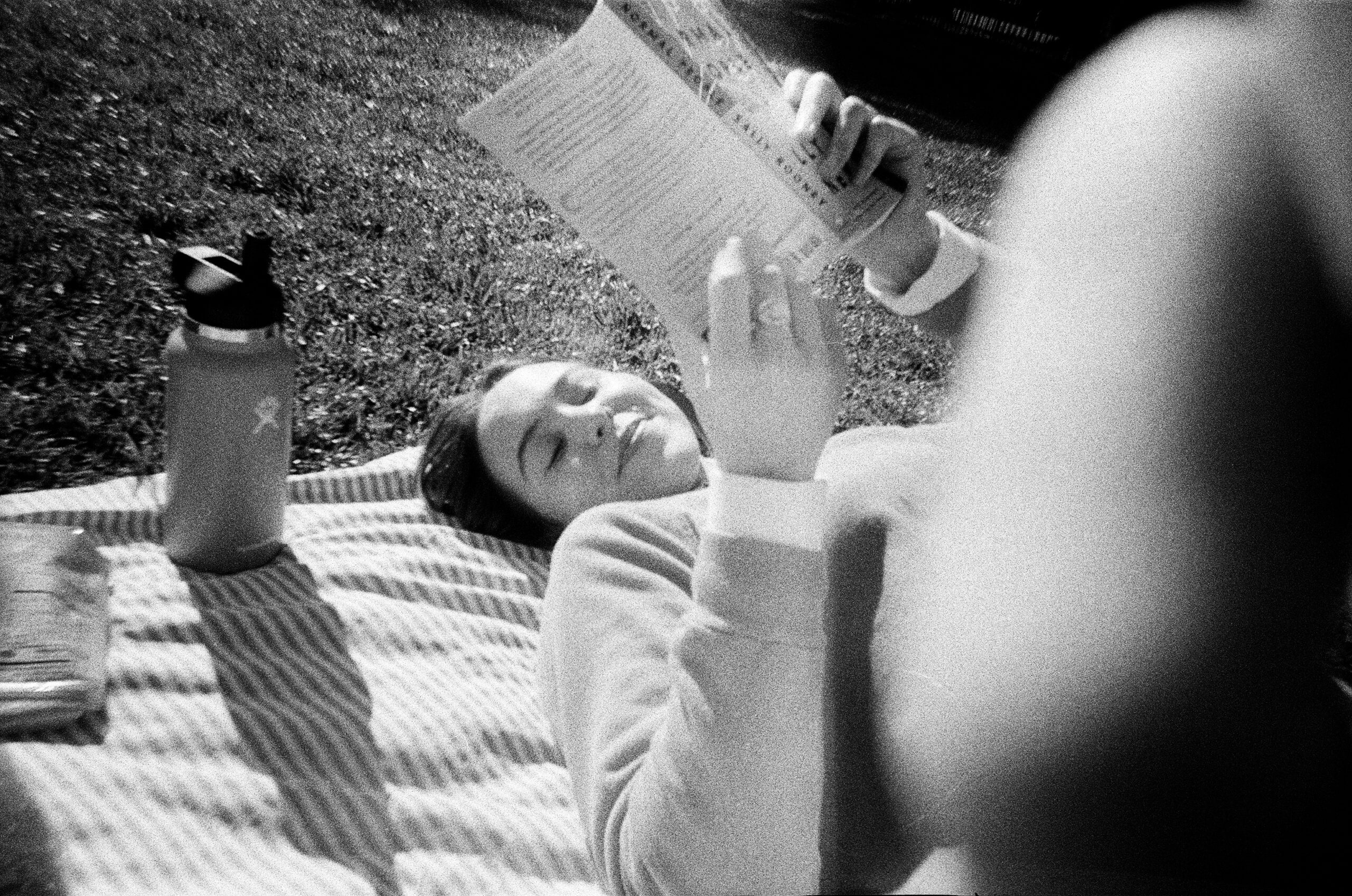 A black, white, and grey photograph of a woman holding a book open while lying on her back on a striped blanket in the grass