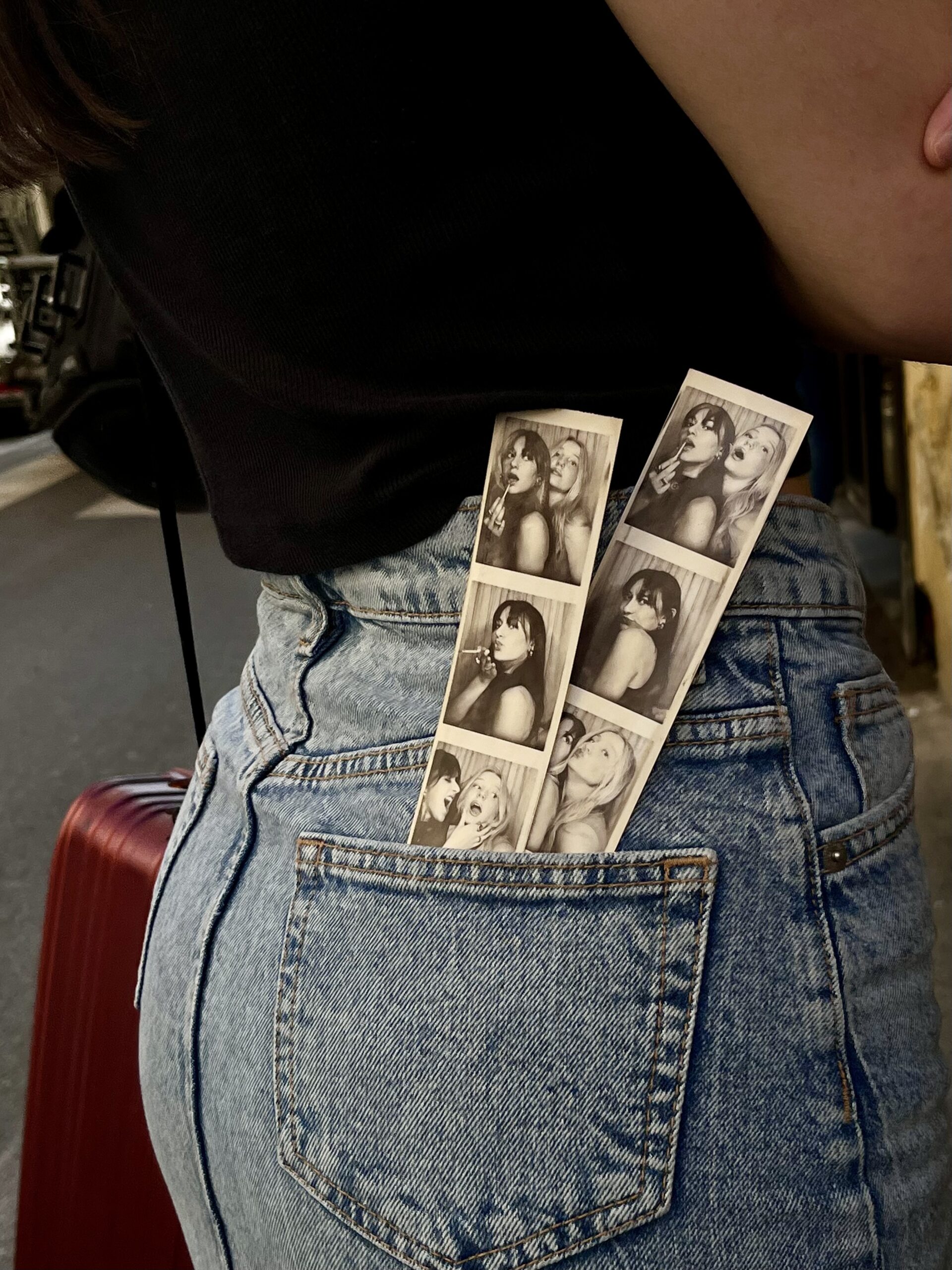 Photos from a Photo Booth stick out from a woman's jean back pocket, with her arms folded across her chest, her black t-shirt partially in view.