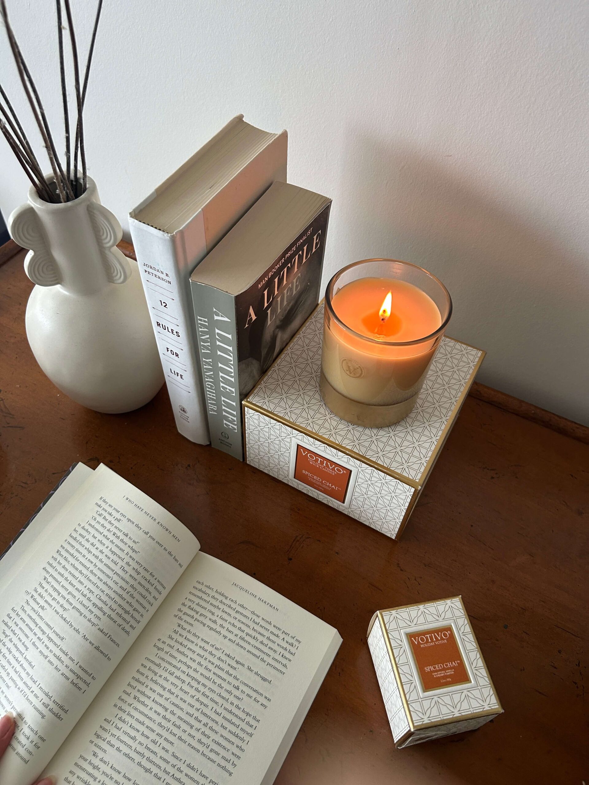 A wooden desk with five books on top of it, one of which is open, a lit candle, and a room diffuser.